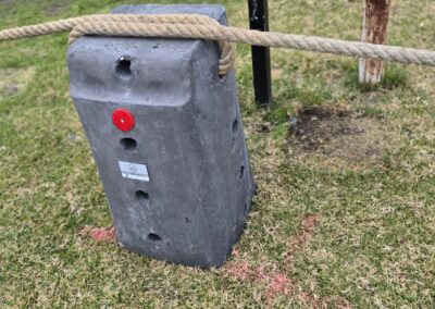 A close-up view of a robust concrete anchor block used to secure a rope barrier in a grassy area. The block features a bright red identification dot and several drilled holes for attaching ropes or cables. The rope is securely looped around the anchor, ensuring stability for the fencing system. The background showcases a well-maintained lawn, emphasizing a neat and organized outdoor space, ideal for settings like cabins, dwellings, caravans, and glamping tents, where outdoor aesthetics and safety are prioritized.