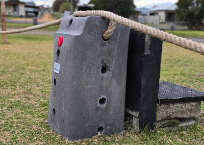 A close-up view of a concrete anchor block secured by a natural fiber rope, featuring a bright red dot for easy identification. The block has several drilled holes designed for attaching the rope, enhancing its functionality in outdoor settings. Nearby, a black support structure can be seen, indicating a sturdy installation. The backdrop showcases a well-maintained grassy area and residential buildings, highlighting its use in creating a secure and private environment around cabins, dwellings, caravans, and glamping tents.