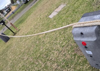 An angled view of two concrete anchor blocks connected by a thick natural fiber rope, demonstrating their role in securing a barrier or fencing system in an outdoor space. Each block features a vibrant red dot for identification, along with multiple drilled holes for attaching the rope. The background reveals a lush green lawn and residential buildings, creating a functional yet aesthetically pleasing environment. This setup is ideal for enhancing privacy and safety around cabins, dwellings, caravans, and glamping tents, showcasing an effective outdoor solution.