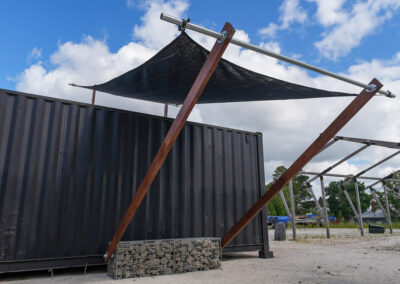 A dynamic view of a shade structure supported by wooden beams, featuring a black shade cloth and a gabion stone base, contributing to the aesthetics of cabins, dwellings, caravans, & glamping tents.