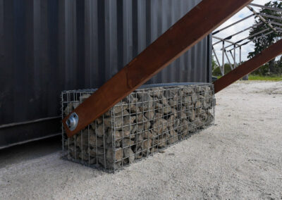 A close-up of a gabion stone support base for a shade structure, emphasizing rustic design elements in a glamping environment, suitable for cabins, dwellings, caravans, & glamping tents.