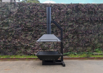 A sleek, modern outdoor barbecue grill designed for communal cooking, with a camouflage background, enhancing the setting for cabins, dwellings, caravans, & glamping tents.