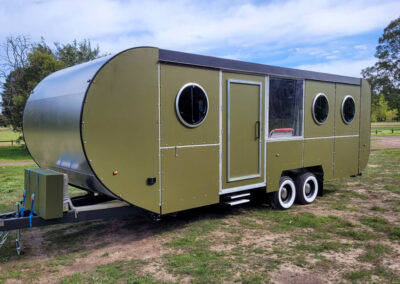 Modern green caravan with a unique rounded shape, circular windows, and double-axle wheels, perfect for Cabins, Dwellings, Caravans, & Glamping Tents. This stylish and mobile accommodation option provides a comfortable, nature-friendly lodging experience, ideal for those seeking adventure and relaxation in scenic outdoor settings