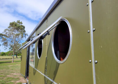 Close-up of a modern green caravan with circular windows, one propped open to allow ventilation, ideal for Cabins, Dwellings, Caravans, & Glamping Tents. The sleek design and functional windows make this caravan a stylish and comfortable lodging option for nature enthusiasts seeking a unique outdoor experience