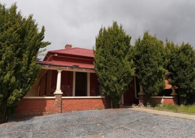 Rustic red-brick building with a red roof, surrounded by tall green trees, featuring a covered porch with columns, ideal for Cabins, Dwellings, Caravans, & Glamping Tents. This charming structure offers a cozy and natural environment, perfect for guests seeking a tranquil escape in a rural setting