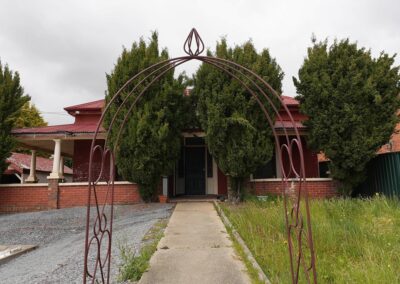 Charming entrance to a rustic red-brick dwelling framed by a decorative metal archway and tall green trees, ideal for Cabins, Dwellings, Caravans, & Glamping Tents. The welcoming pathway leads to a covered porch, offering a serene and inviting atmosphere perfect for guests seeking a tranquil retreat in nature