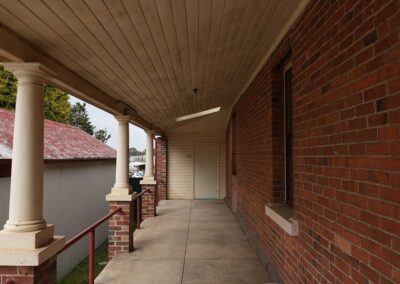 Long, covered porch with columns and red brick walls, offering a rustic outdoor walkway for guests in Cabins, Dwellings, Caravans, & Glamping Tents. This inviting space provides a sheltered area to relax and enjoy the surrounding natural scenery, enhancing the charm of rural accommodations