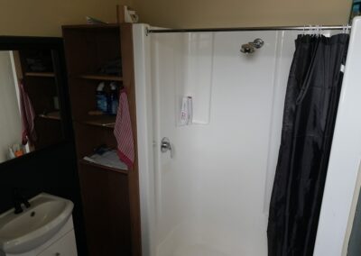 Compact bathroom with a white shower stall, black shower curtain, small sink, and storage shelves holding cleaning supplies and towels, ideal for guests in Cabins, Dwellings, Caravans, & Glamping Tents. This practical and organized setup offers essential amenities for a comfortable stay in rustic or glamping accommodations