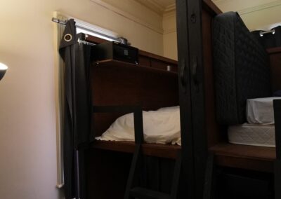 Cozy bunk bed setup with wooden frame, ladder access, and bedding, designed for guests in Cabins, Dwellings, Caravans, & Glamping Tents. This compact sleeping arrangement includes privacy curtains and a small shelf with a safe, providing a practical and secure sleeping space for travelers seeking comfort and convenience